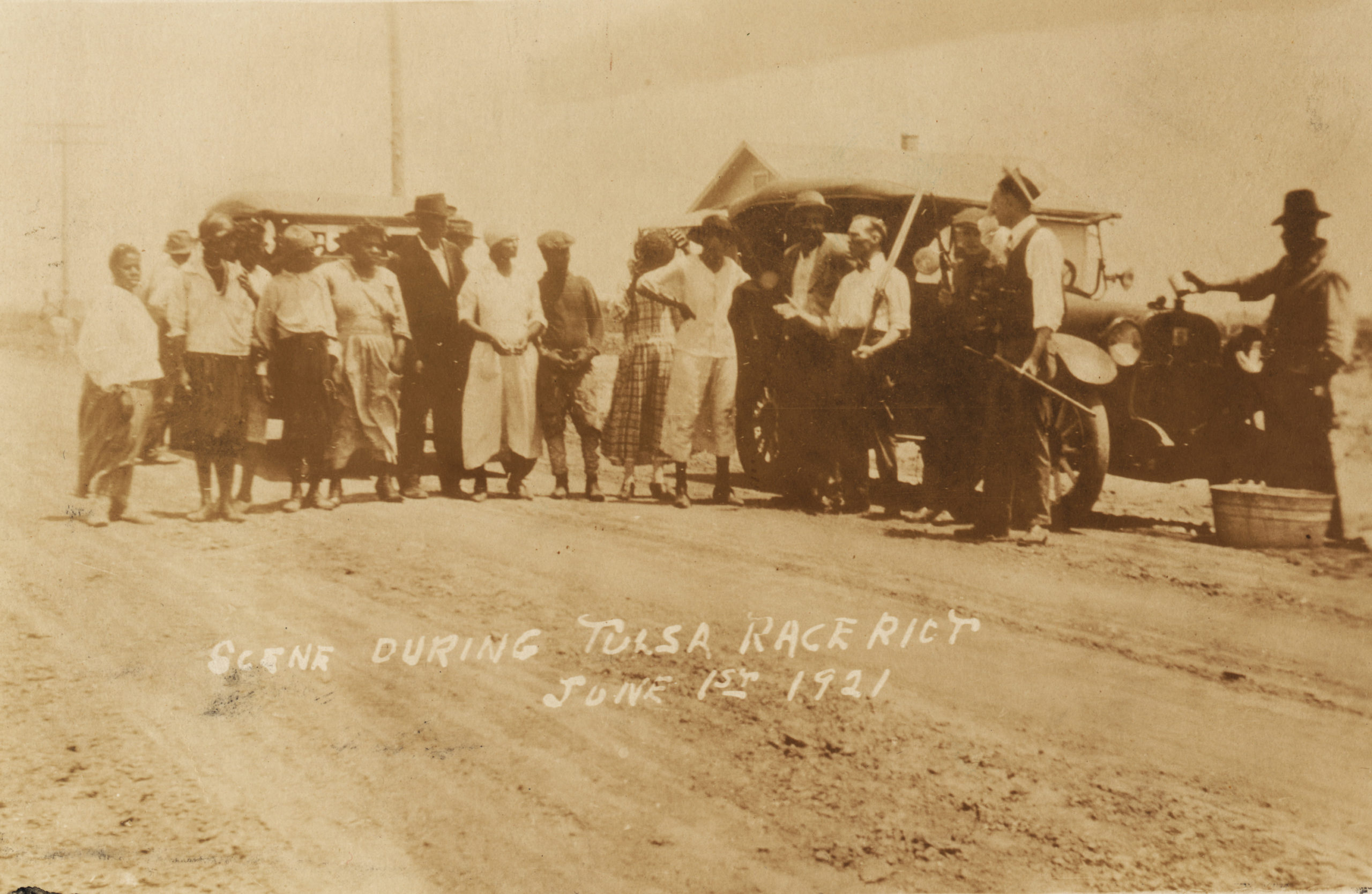 A Postcard showing a group of African-Americans and armed Whites in civilian attire standing on a road. There is a building and two vehicles behind the crowd. One man is refilling the radiator of one of the vehicles. Title is taken from the writing on the face of the postcard.