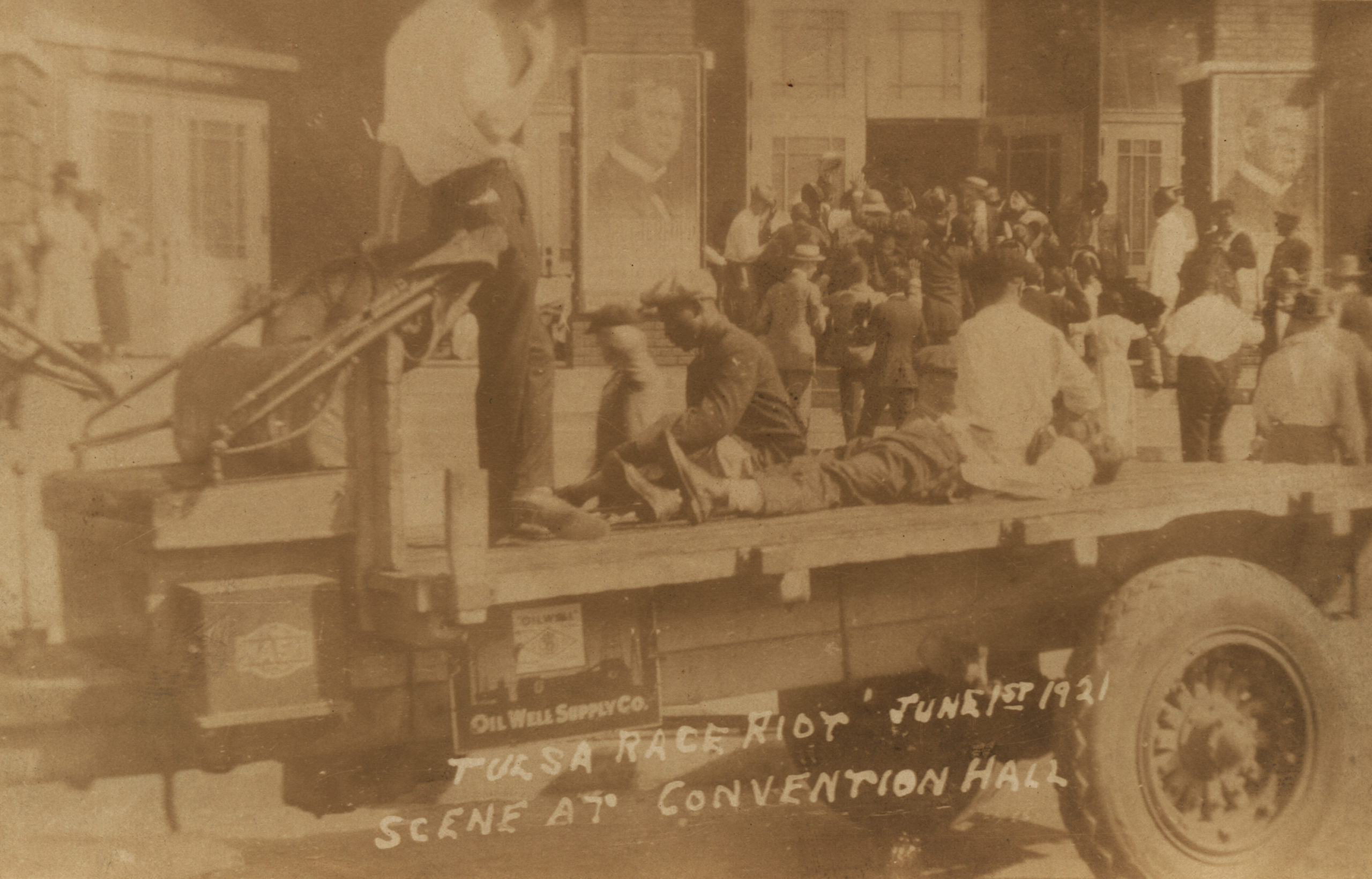 A postcard showing a truck parked in front of the Convention Hall. One man lies on the bed of the truck, either wounded or dead, while two others sit to either side. A man in civilian attire stands guard over them.