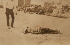 A postcard showing a man lying dead in the street, with a sheet, or piece of paper covering his face. This victim appears in a number of photographs in this collection, taken from different angles at different times of the morning.