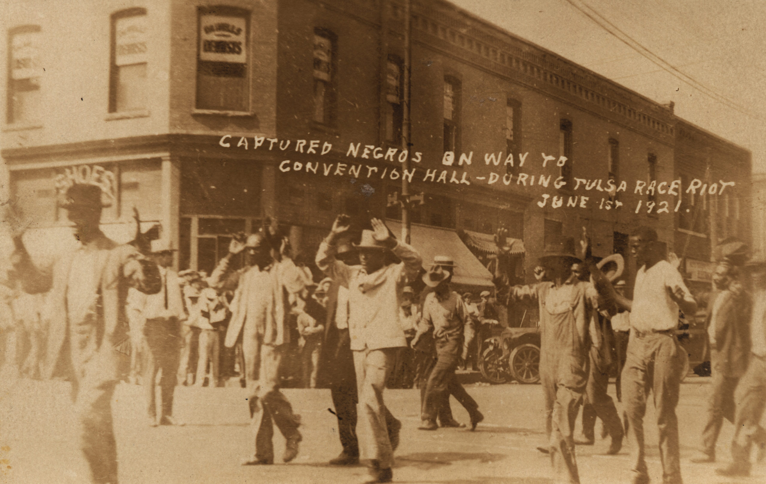 A postcard showing a group of detainees being marched past the corner of 2nd and Main under armed guard. The building in the background is 202 S. Main, on the southwest corner. Based on the shadows of the building and the people, it is late morning. They are heading east (or are turning to head east) on 2nd, so it is more likely that they are among those being marched south towards McNulty Park than to be heading towards the Convention Hall, which is several blocks north of this intersection.