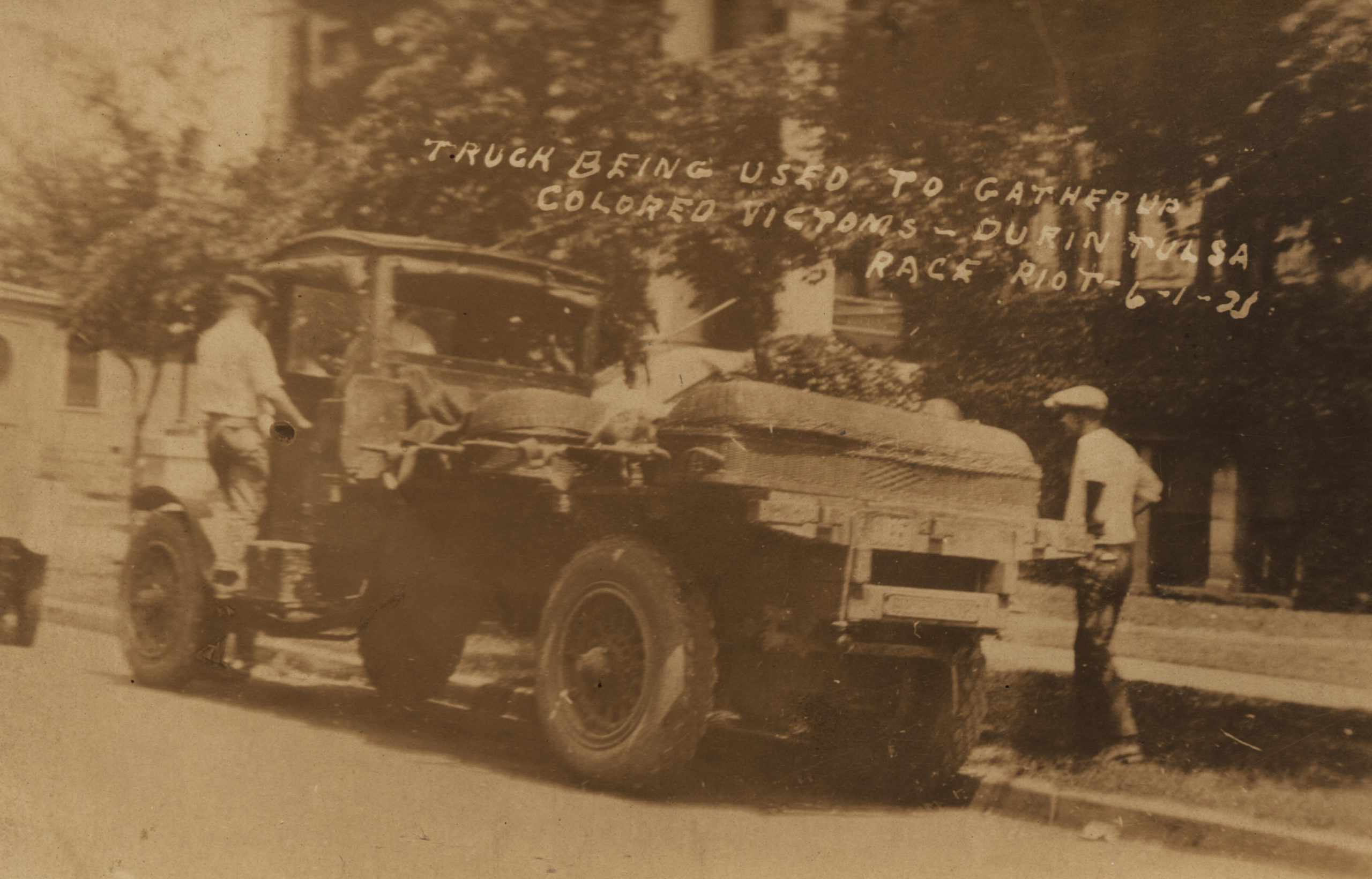 A Postcard showing a pair of men who have loaded two wicker coffins onto a truck at the Courthouse. Title is taken from the writing on the face of the Postcard.