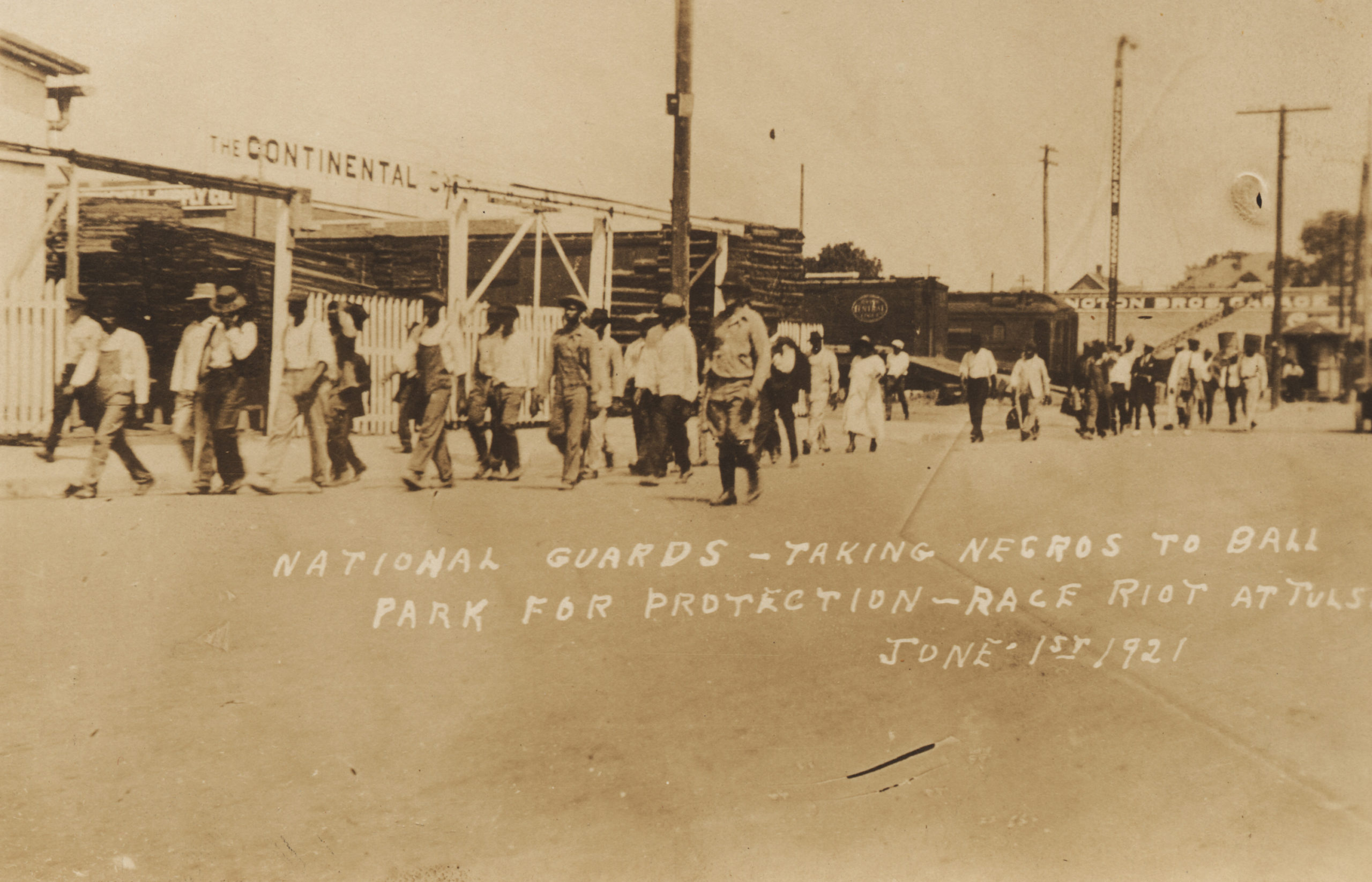 A postcard made from a photograph (1989.004.5.58). A large group of people are being escorted by several men in civilian attire with an automobile alongside. They have just crossed the tracks and are passing in front of the Continental Supply Co. (offices at 19 S. Main). There is an issue with this image since while the Continental Supply Co. on the south side of the tracks, the address is on the east side of the street. The structures in the rest of the image are also not consistant with this being that part of Main St. Title is taken from the writing on the face of the postcard.