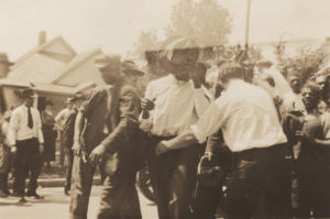 Photographic print of African-American men being detained and led down a residential street. White men and women stand in the background.