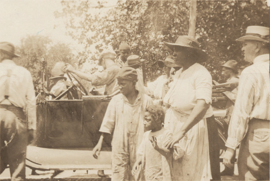 Photographic print of several people standing near a truck, most particularly a woman with a boy and young girl.
