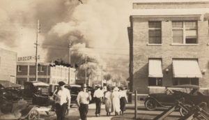 A photographic print showing the scene looking up Elgin Ave. from the alley between 1st St. and 2nd St. The Building on the right is the Santa Fe Freight office. Two armed men in civilian attire are walking away from the riot area, while a man and three women walk towards it. In the distance a crowd has gathered. “32” printed on back. “Negro Riot, June 1 1921” written on back. Photos 24-28 are from the same development batch, and notes are in the same handwriting. Digital image has been modified from the original for clarity.