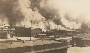 A photographic print showing the burning along Archer the morning of the 1st. This photo was taken north across Elgin Ave. and the Frisco tracks towards Greenwood Ave. (From Dreyfus Bros. Co. on the left to Goodner-Malone on the right at Frankfurt). "32" printed on back. "Riot Tulsa, June 1 1921" written on back. Photos 24-28 are from the same development batch, and notes are in the same handwriting