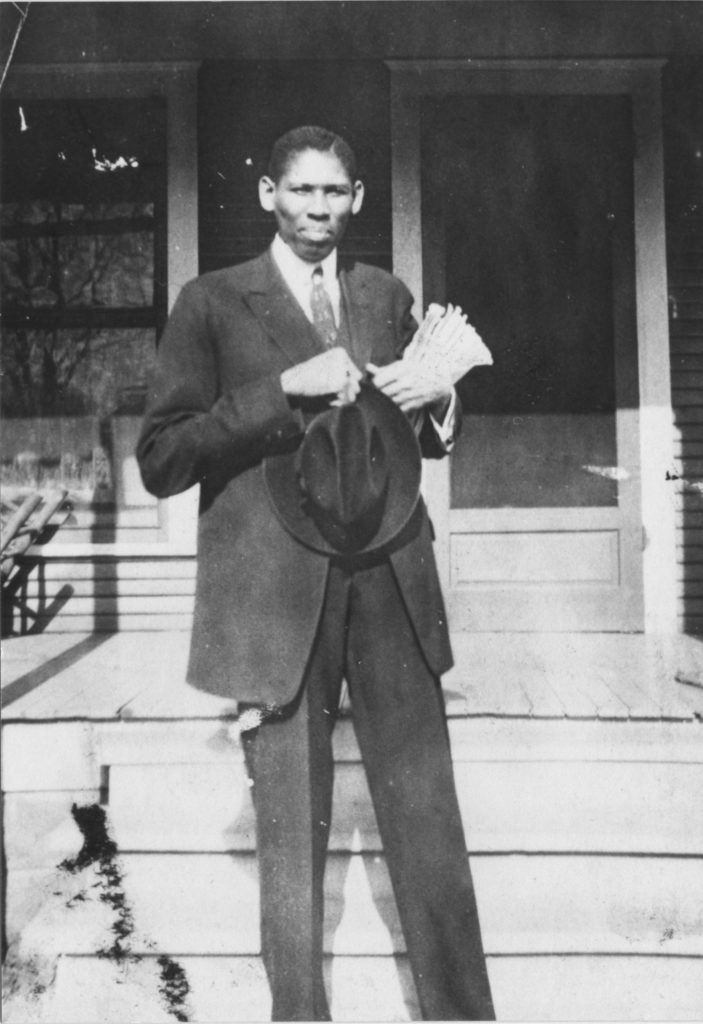 A photographic print depicting James T. A. West, an instructor at Booker T. Washington High School, standing outside a house, holding his hat. This man also appears in photo 1989-004-5-31.