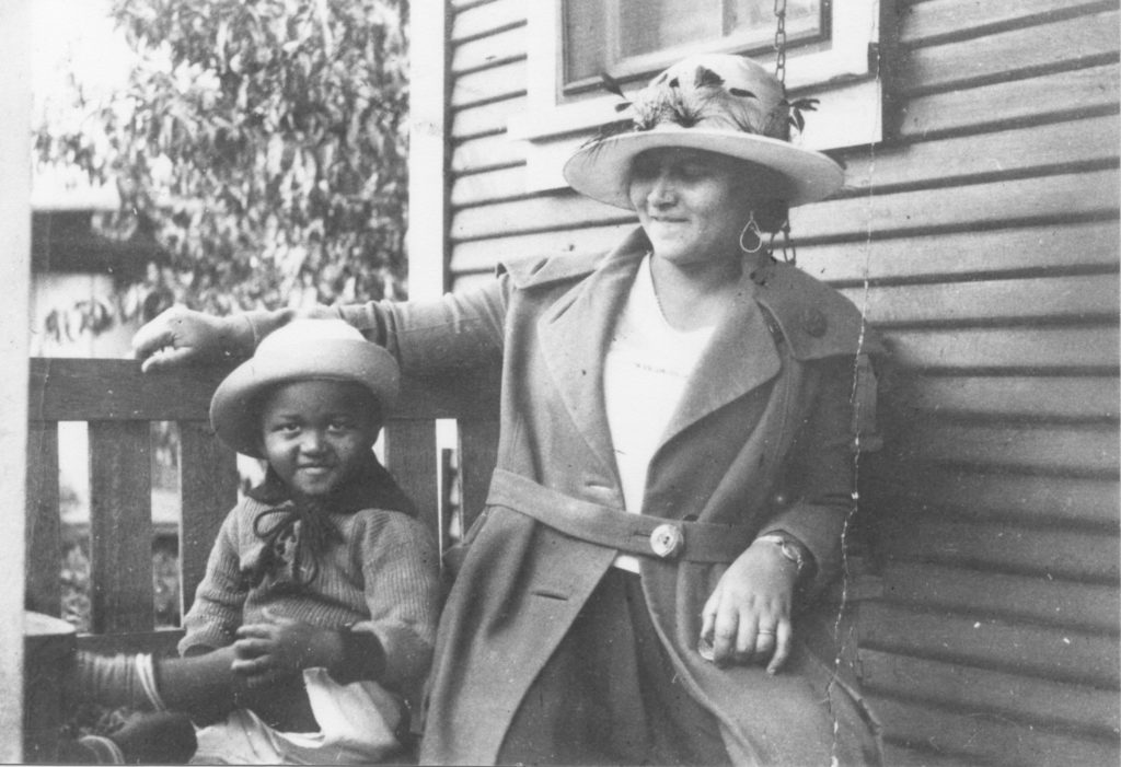 African-American woman and girl sitting on a porch swing, both dressed in coats and hats, by the side of a house.