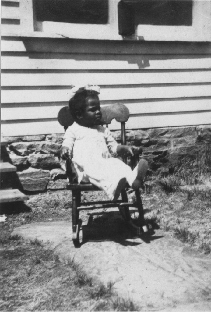 Photograph of a young African-American girl wearing a white dress, shoes, and hairbow, sitting in a rocking chair in front of steps leading up to a house.