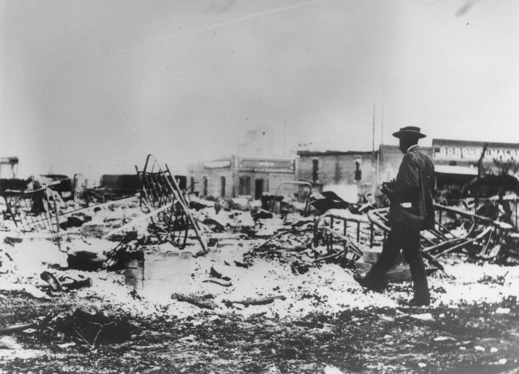 A photographic reproduction of a photo depicting an African-American photographer wearing his safe conduct badge looking at the ruins of the Midway Hotel. The Goodner-Malone Co. is in the background. "OHS:15583B" written on back.