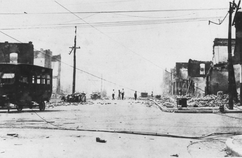A photographic reproduction of a photo taken looking up Greenwood Ave. from Archer St. Based on the burnt out cars, the cables, and relatively intact walls (which have not yet collapsed), this photo was taken fairly early in the cleanup. "G5 of 28" written on back.
