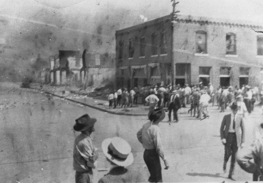 A photographic reproduction of a photo taken of Greenwood Ave. and Archer St. Note that while the building on the left (Woods Bldg. 101-107 N. Greenwood) has not yet been burned and is being looted, the rest of the east side of Greenwood has been burned. "G17 of 28" written on back. Title written on front. Photographer has not been identified.