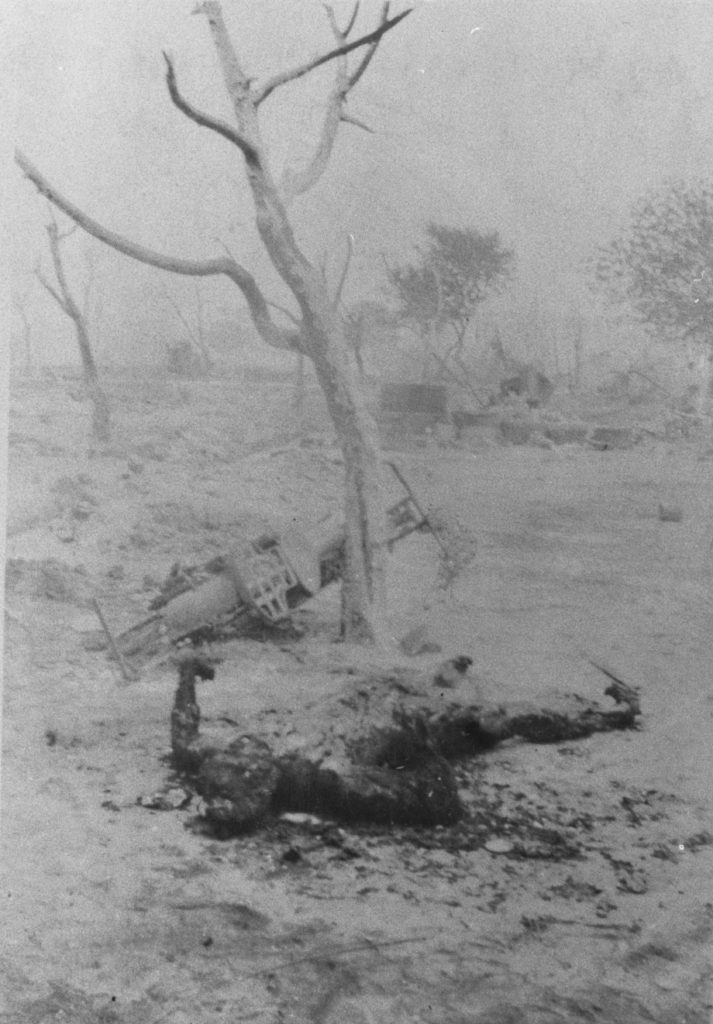 A photographic reproduction of a photo taken of burned corpse. A burned motorcycle lies against a tree. This body appears in photos 1989.004.5.14, 1989.004.5.55, and 1989.004.5.S10. "G21 of 28" written on back (struck out). Title written on front.