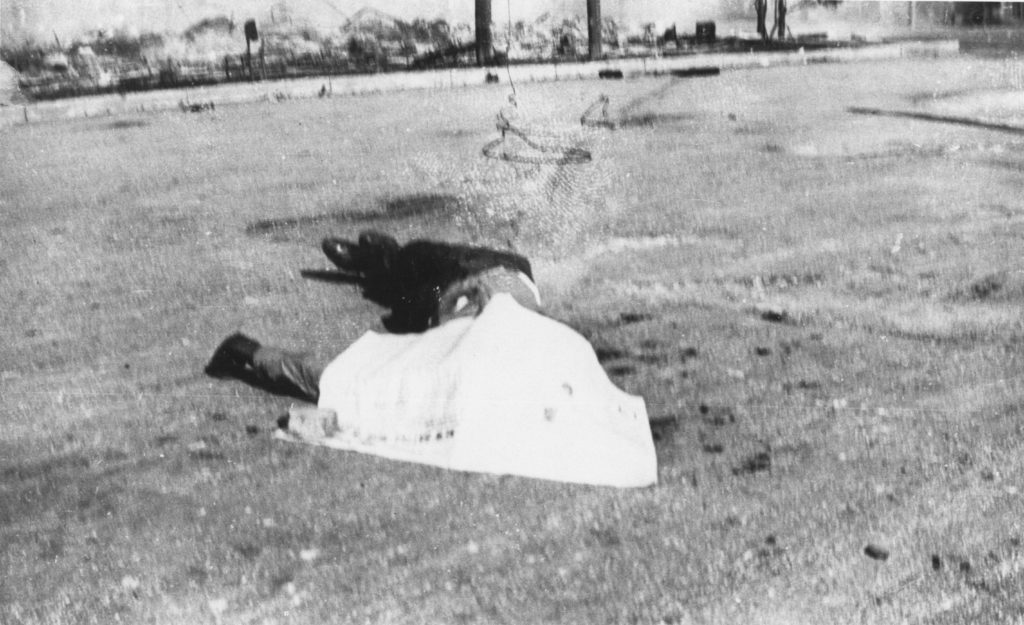 A photographic reproduction of a photo taken of corpse in the street with a sheet, or piece of paper covering his face. This body appears in photos 1989.004.5.6, 1989.004.5.57, and 1989.004.5.W18. "G23 of 28" written on back (struck out). Title written on front.