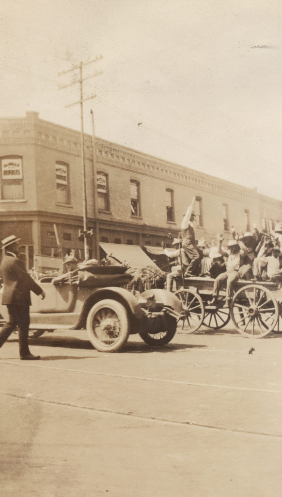 A small group of people are held on the back of a mule drawn wagon, with their hands held up. An automobile is driving between the wagon and the photographer, two men in the car in civilian attire, one of whom has a rifle with a mounted bayonet. The building in the background is 202 S. Main, on the southwest corner of 2nd and Main. Based on the shadows of the building and the people, it is late morning.